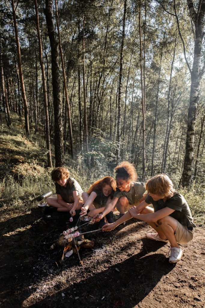 Group of teens roasting marshmallows over a campfire in a sunlit forest setting.